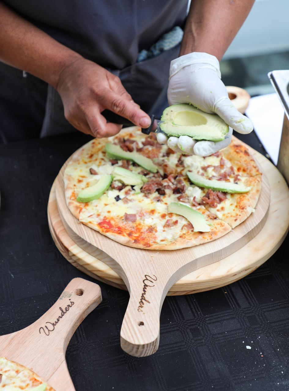 Fresh avocado being sliced onto a bacon and feta pizza on a wooden board