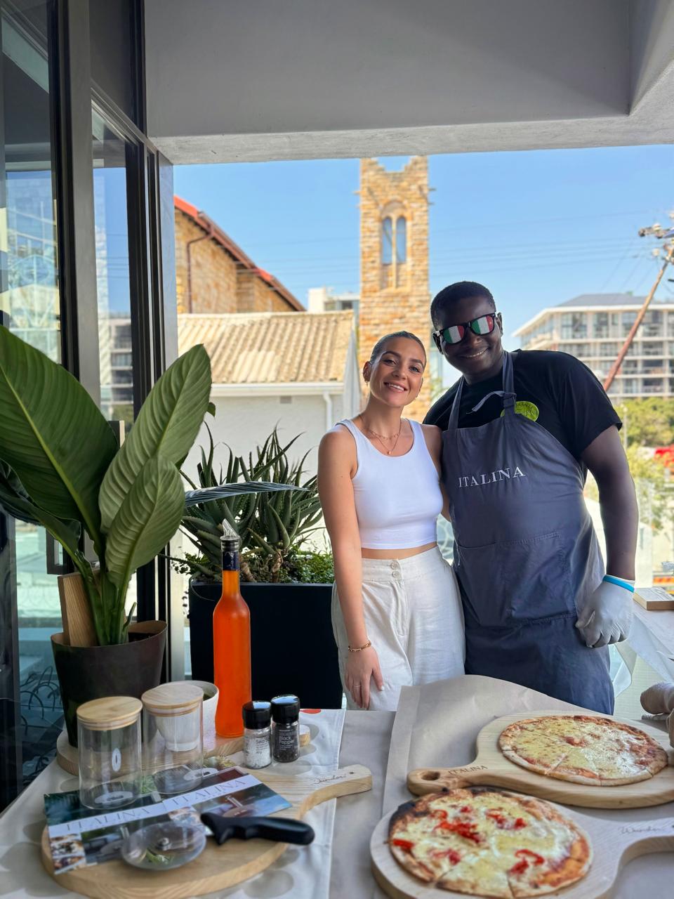 Hosts and Italina pizzaiolo behind the pizza setup on a sunny balcony