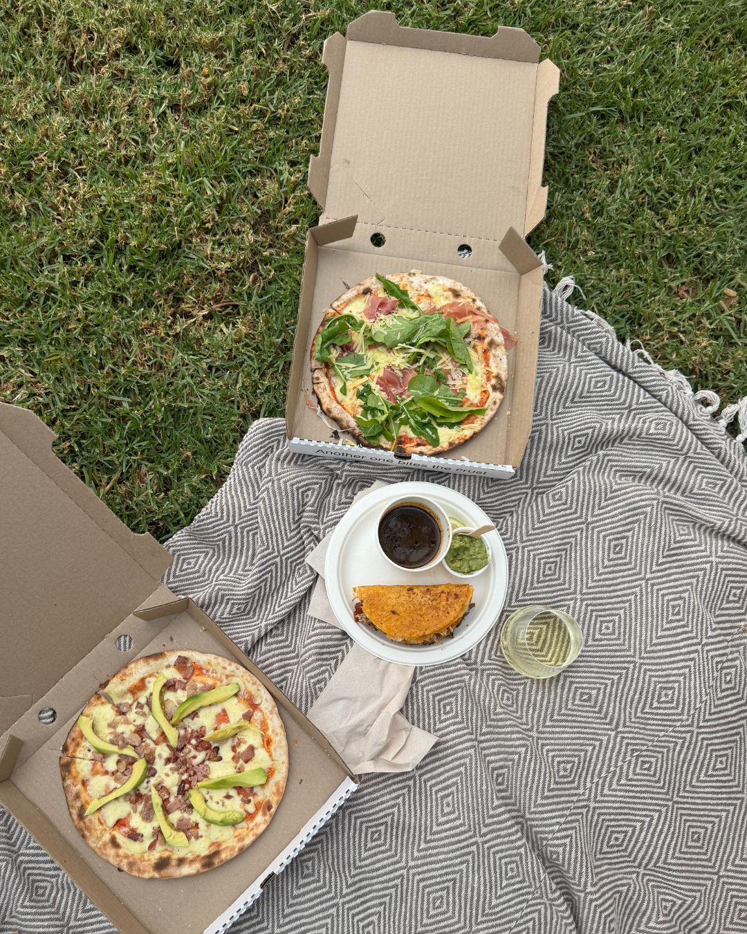 Two boxed pizzas on a picnic blanket with dips and wine
