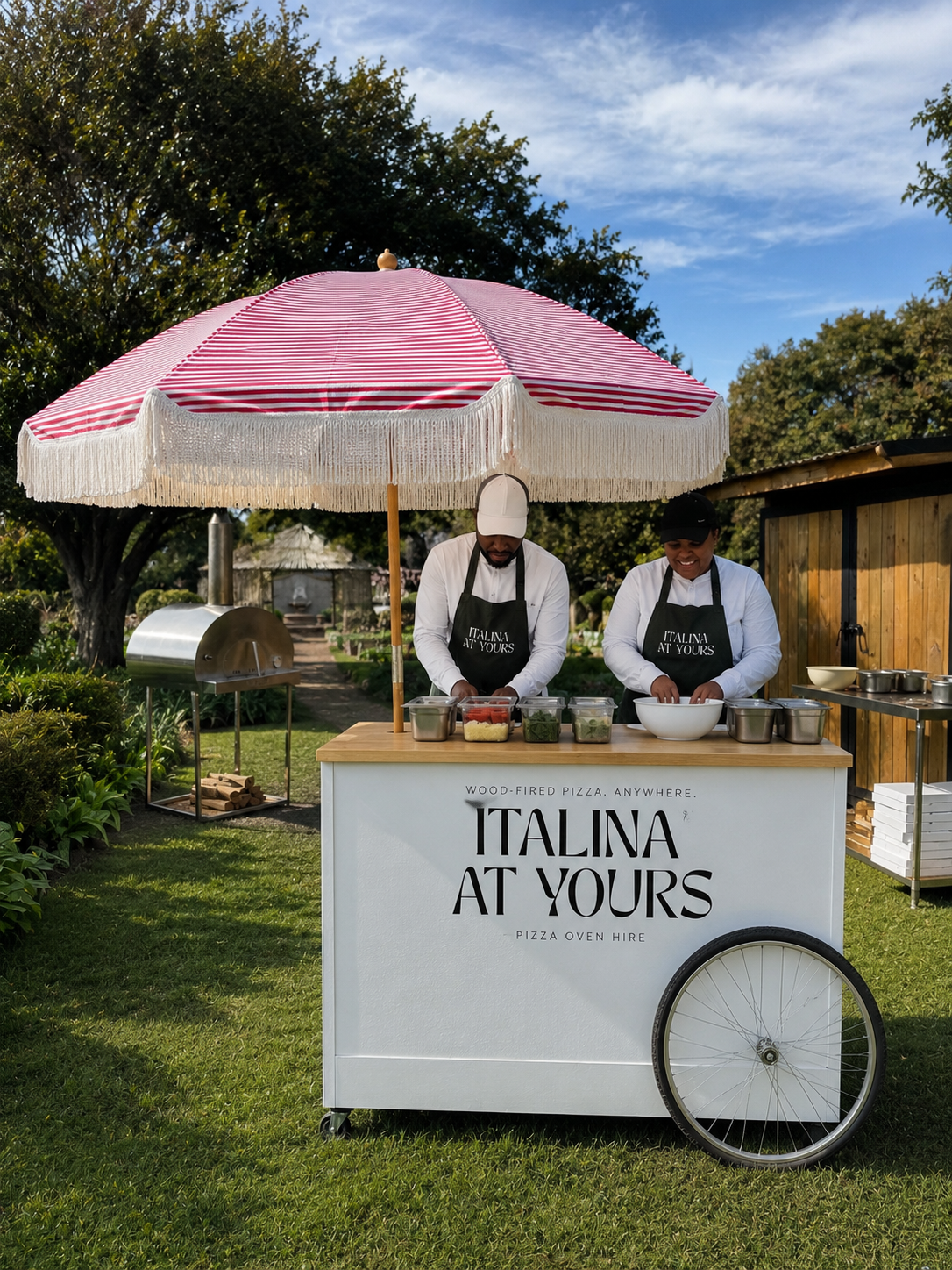 The Italina At Yours pizza cart with a fringed umbrella and crew prepping toppings on a lawn