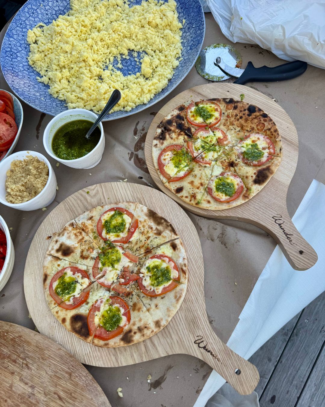 Two tomato and pesto pizzas on wooden paddles with toppings alongside